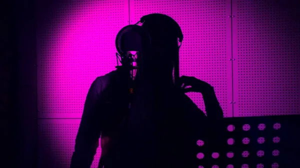 Μια νεαρή γυναίκα τραγουδάει ένα τραγούδι σε ένα μικρόφωνο σε ένα στούντιο ηχογραφήσεων κάτω από φως νέον — Φωτογραφία Αρχείου