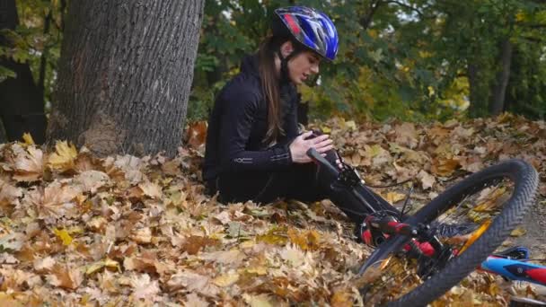 En ung kvinna sitter i en park efter att ha fallit från en cykel, lugnar smärta i knäet, är en farlig cykeltur i parken en höstdag — Stockvideo