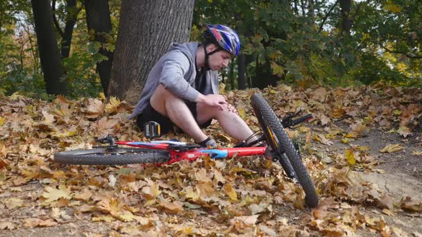 Un joven se sienta en un parque después de caer de una bicicleta, calma el dolor en la rodilla, es un peligroso paseo en bicicleta en el parque en un día de otoño — Vídeo de stock