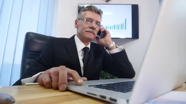 Hombre mayor con teléfono inteligente blanco habla y sonríe en la oficina — Vídeo de stock