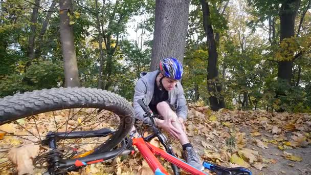 Een jongeman in een park zit na het vallen van een fiets, kalmeert de pijn in zijn knie, is een gevaarlijke fiets rijden in het park op een herfstdag — Stockvideo