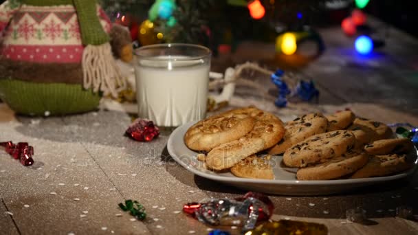 Tavolo marrone in legno decorato con roba natalizia e ghirlande. Un bicchiere di latte e un piatto di biscotti su un tavolo di Natale — Video Stock