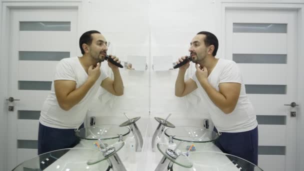 Молодой человек бреет бороду электробритвой в ванной комнате — стоковое видео