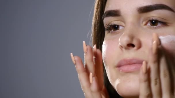 Joven hermosa mujer aplicando crema en su cara, aislado en gris — Vídeo de stock