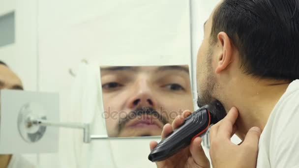 Молодой человек бреет бороду электробритвой в ванной комнате — стоковое видео