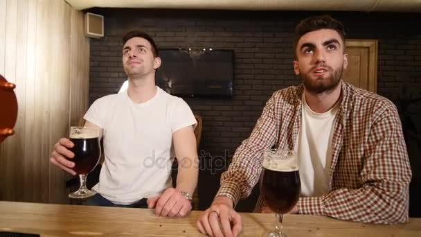 Dos amigos caucásicos celebrando el éxito de su equipo deportivo en el pub. Atractivos jóvenes abrazándose en el mostrador del bar. Aficionados al deporte contentos tintineando sus vasos con cerveza — Vídeos de Stock