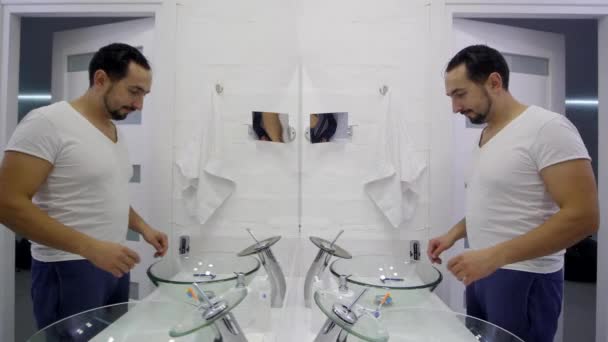 Человек умывается перед зеркалом. Уставший небритый парень в белой футболке — стоковое видео