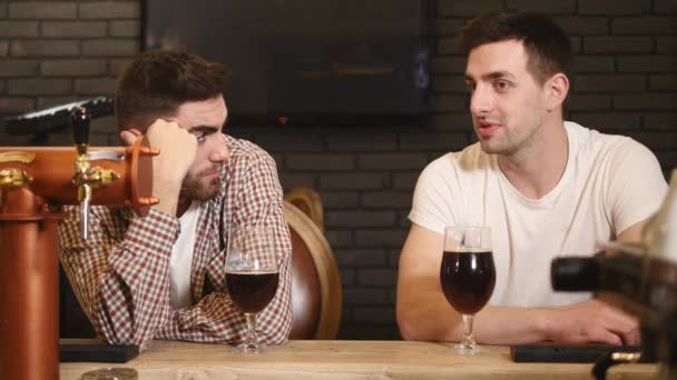 Dos jóvenes amigos sentados en el bar y hablando entre sí mientras beben cerveza — Vídeos de Stock