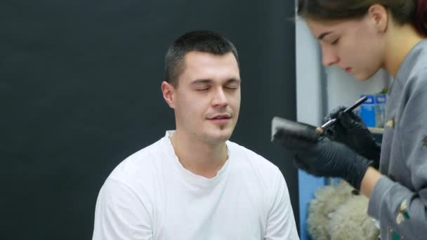 Het meisje maakt aan de man. een professionele make-up artiest make-up een wenkbrauw — Stockvideo