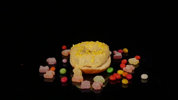 Gelbe lustige überraschte glasierte Donut mit Streusel, gestreifte Karamellbonbons, bunte Dragee mit Rosinen oder Erdnüssen im Inneren drehen sich auf schwarzem Hintergrund — Stockfoto