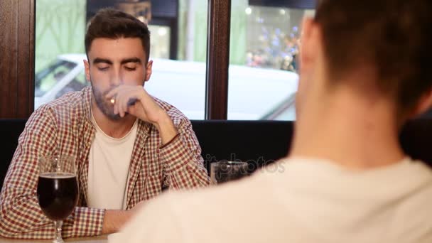 Двоє молодих друзів сидять у барі і розмовляють один з одним під час пиття пива — стокове відео