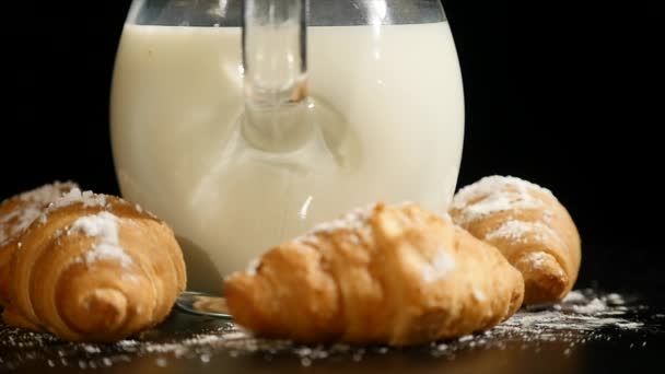 Jarra de leche con croissant girar sobre un fondo negro — Vídeos de Stock