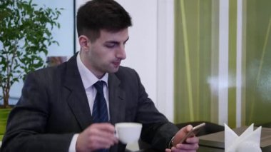 Bir adam kafede bir masada oturur ve telefon kullanır. Sol elinde telefon