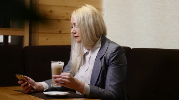 Женщина с помощью приложения на смартфоне, пьет кофе, улыбается, пишет смс на мобильный телефон — стоковое видео