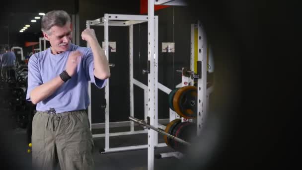 Hombre mayor muestra sus propios bíceps de brazo en el fondo del gimnasio — Vídeo de stock
