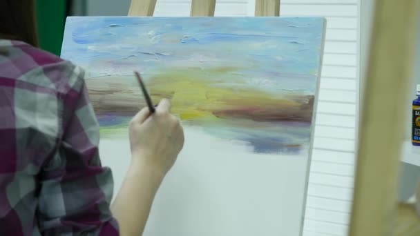 Μια νεαρή γυναίκα ζωγραφίζει έναν πίνακα ζωγραφικής σε καμβά, στέκεται πίσω από ένα καβαλέτο, σε ένα ατμοσφαιρικό εργαστήρι — Αρχείο Βίντεο
