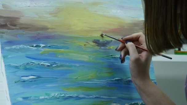 Een jonge vrouw schildert een schilderij op een canvas, staande achter een schildersezel, in een sfeervolle atelier — Stockvideo