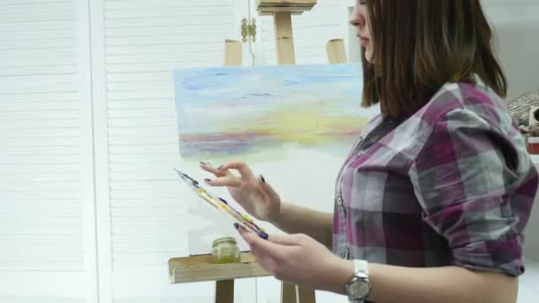 Μια νεαρή γυναίκα ζωγραφίζει έναν πίνακα ζωγραφικής σε καμβά, στέκεται πίσω από ένα καβαλέτο, σε ένα ατμοσφαιρικό εργαστήρι — Αρχείο Βίντεο