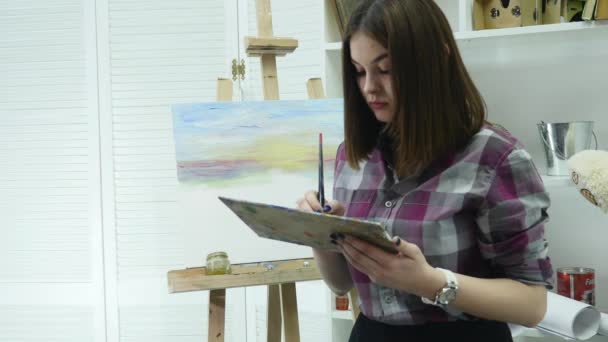 Een jonge vrouw schildert een schilderij op een canvas, staande achter een schildersezel, in een sfeervolle atelier — Stockvideo