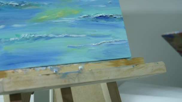 Una joven pinta una pintura sobre un lienzo, de pie detrás de un caballete, en un taller atmosférico — Vídeo de stock