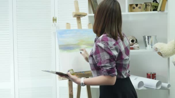 Ung kvinnelig kunstner i forkle maler bilde på lerret i kunststudioet – stockvideo