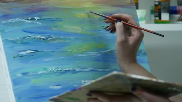 Una joven pinta una pintura sobre un lienzo, de pie detrás de un caballete, en un taller atmosférico — Vídeos de Stock