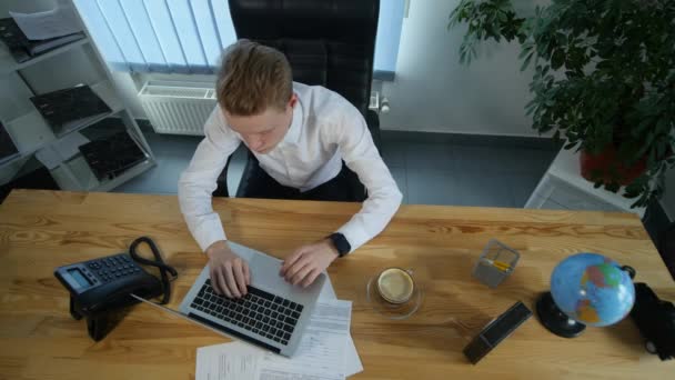 Geschäftsmann, der am Schreibtisch mit seinem Laptop arbeitet und Kaffee trinkt, umgeben von viel Papierkram und Finanzberichten — Stockvideo