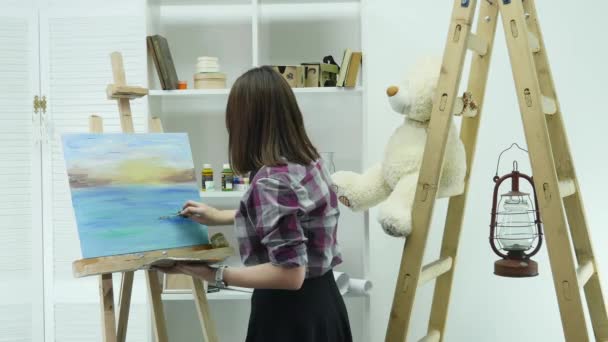 O tânără pictează o pictură pe o pânză, stând în spatele unui șevalet, într-un atelier atmosferic. — Videoclip de stoc