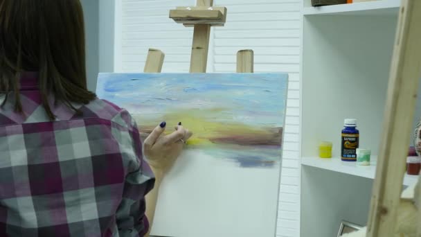 Kunstneren maler en kvinne på lerretet og smører en bred pensel. Lerretet står på staffeliet. Kunstneren trekker mot staffeliet – stockvideo