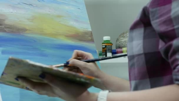 Una joven pinta una pintura sobre un lienzo, de pie detrás de un caballete, en un taller atmosférico — Vídeo de stock