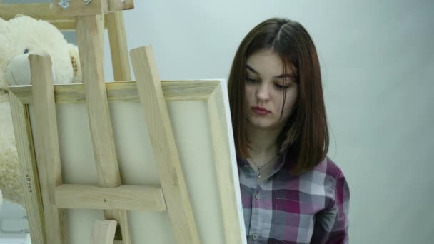 Молодая женщина рисует картину на холсте, стоящем за мольбертом, в атмосфере мастерской — стоковое видео