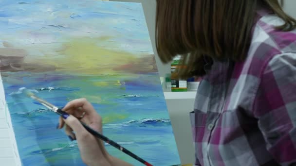 A young woman paints a painting on a canvas, standing behind an easel, in an atmospheric workshop — Stock Video