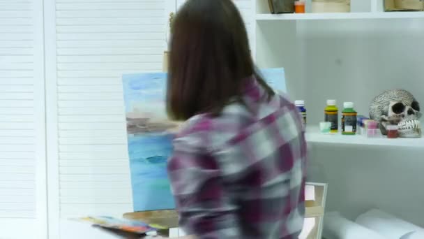 A young woman paints a painting on a canvas, standing behind an easel, in an atmospheric workshop. time lipse — Stock Video