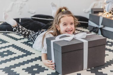 Uykulu ama mutlu bir çocuk Noel pijamalarıyla yerde yatıyor, Noel ağacının altında, çenesini büyük hediyeye uzatıyor..