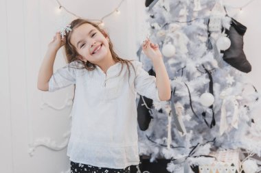 Gülümseyen kız, küçük kar prensesi, Noel ağacının yanında, beyaz bir arka planda..