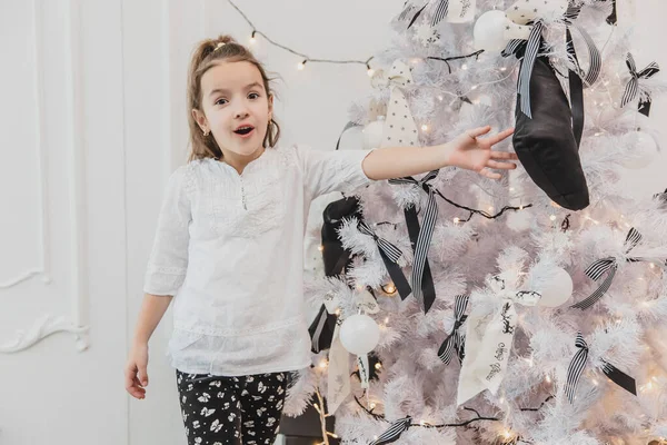 Aufgeregtes kleines Mädchen mit offenem Mund zeigt auf den wunderschön geschmückten Weihnachtsbaum mit Lichtern, Kugeln und Socken. — Stockfoto