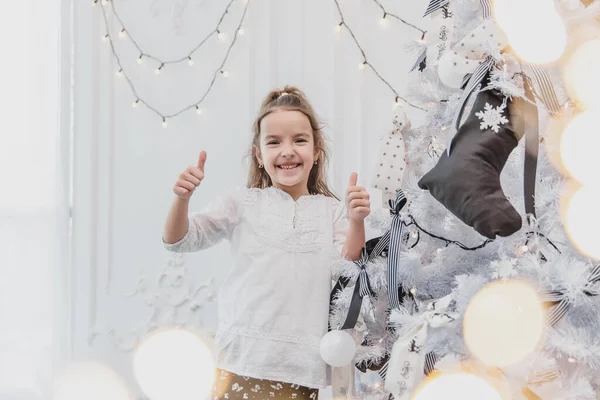 Kleines Kind mit lustigem Pferdeschwanz steht neben Weihnachtsbaum und zeigt Daumen hoch, weil ihr Baum der beste ist. — Stockfoto