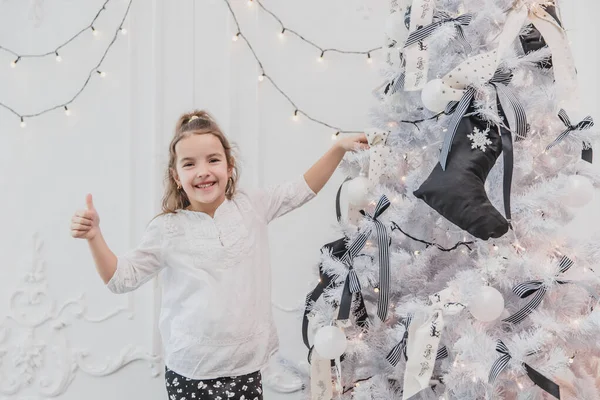 Маленький ребенок со смешным хвостиком стоит возле рождественской елки, показывая большие пальцы вверх, потому что ее дерево является лучшим . — стоковое фото