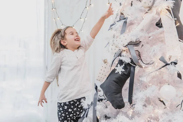 Возбужденная маленькая девочка указывает на красиво украшенную елку с огнями, шарами и носками . — стоковое фото