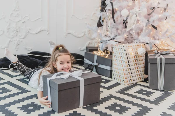 Сонный, но счастливый ребенок в рождественской пижаме лежит на полу под елкой, держа подбородок на большом подарке . — стоковое фото