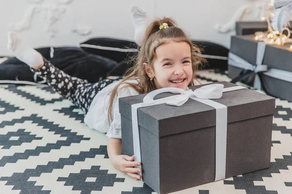 Un niño soñoliento pero feliz en pijama de navidad yace en el suelo bajo el árbol de navidad, manteniendo su barbilla en el gran regalo . — Foto de Stock