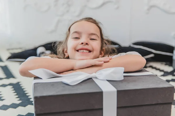 Primer plano de la hermosa niña ángel con una sonrisa perfecta que yace en el suelo, manteniendo las manos en la caja de regalo grande con cinta . — Foto de Stock