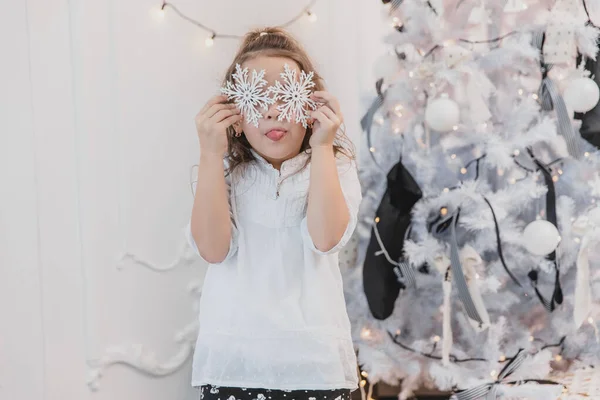 Bambina che gioca con i fiocchi di neve dell'albero di Natale, nascondendosi dietro di loro e facendo smorfie . — Foto Stock