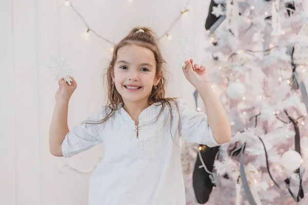 Kleines brünettes Mädchen schmückt zu Hause einen Weihnachtsbaum mit silbernen Schneeflocken. — Stockfoto