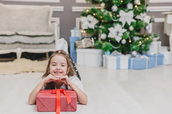 Gros plan de belle petite fille ange avec un sourire parfait posé sur le sol, gardant les mains sur une grande boîte cadeau avec ruban . — Photo