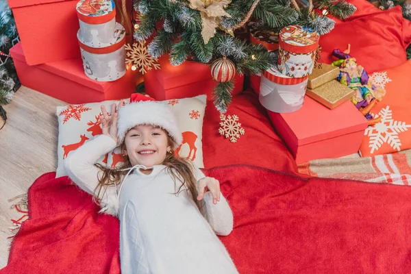 Weihnachtsmann-Kind liegt auf dem Kopfkissen mit Rentier-Print und roter Decke unter dem Weihnachtsbaum. — Stockfoto