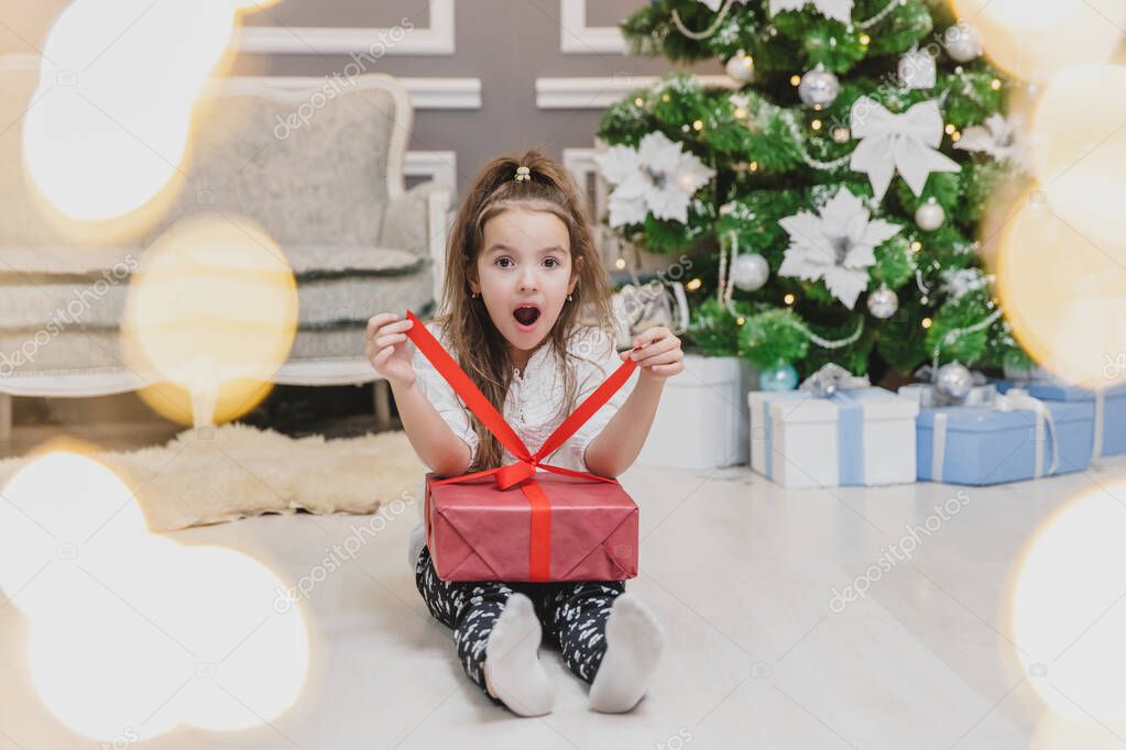 Full-body, forefront blurred photo of cute child sitting in lotos position with a Christmas present in hands, finding out what is inside.