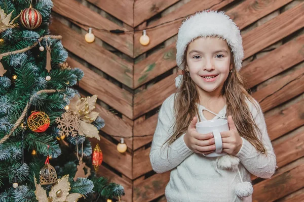 Photo en gros plan de mignonne fille souriante assise confortablement à la maison, tenant une tasse de chocolat chaud à l'arbre de Noël sur fond en bois . — Photo