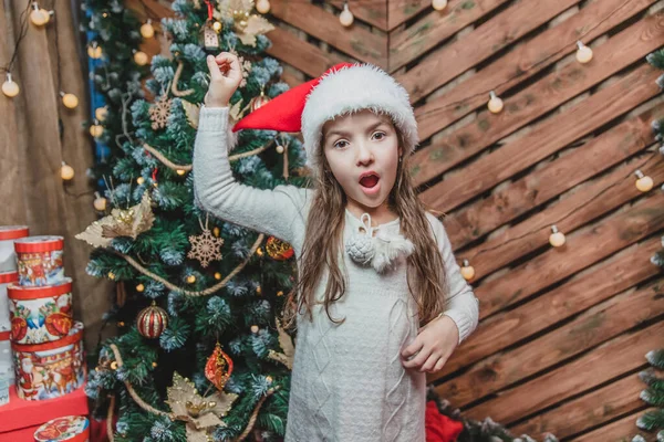Bambino divertente vicino abete, tirando su il cappello di Babbo Natale e guardando la fotocamera in previsione di un miracolo di Natale . — Foto Stock