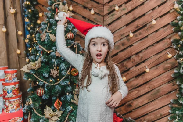 Bambino divertente vicino abete, tirando su il cappello di Babbo Natale e guardando la fotocamera in previsione di un miracolo di Natale . — Foto Stock
